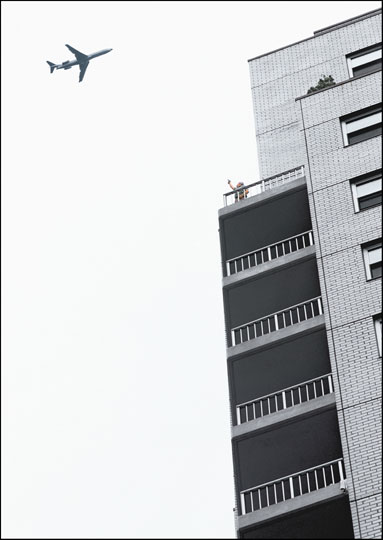 A 747 jet flies low and close to the top of an apartment tower. On a balcony, a person poses with their arm in the air, pointing a gun at the plane.